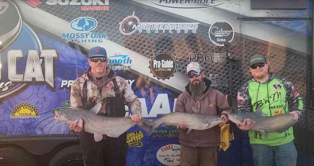 Spencer Bauer and two fellow anglers stand in front of a tournament trailer, each holding a large catfish.
